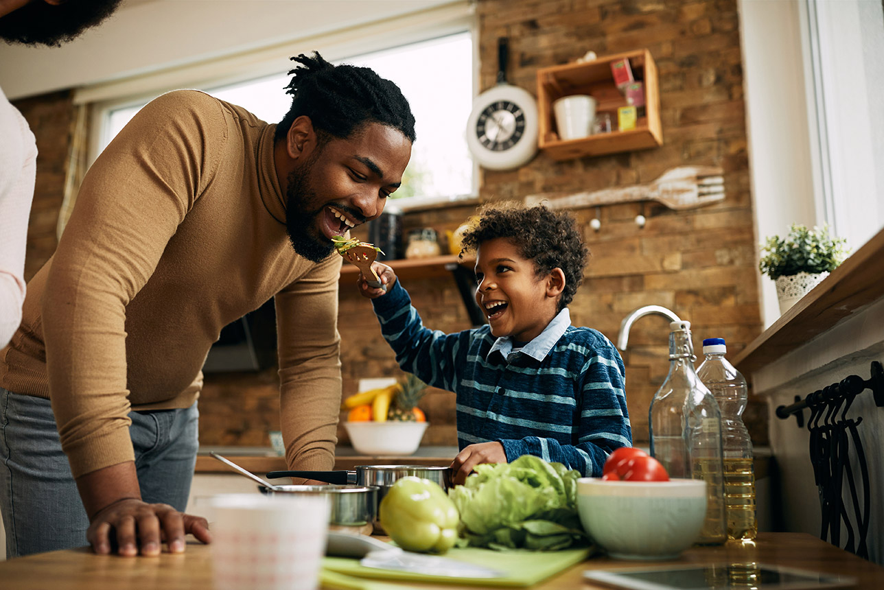 Happy African American boy having fun while feeding his father in the kitchen