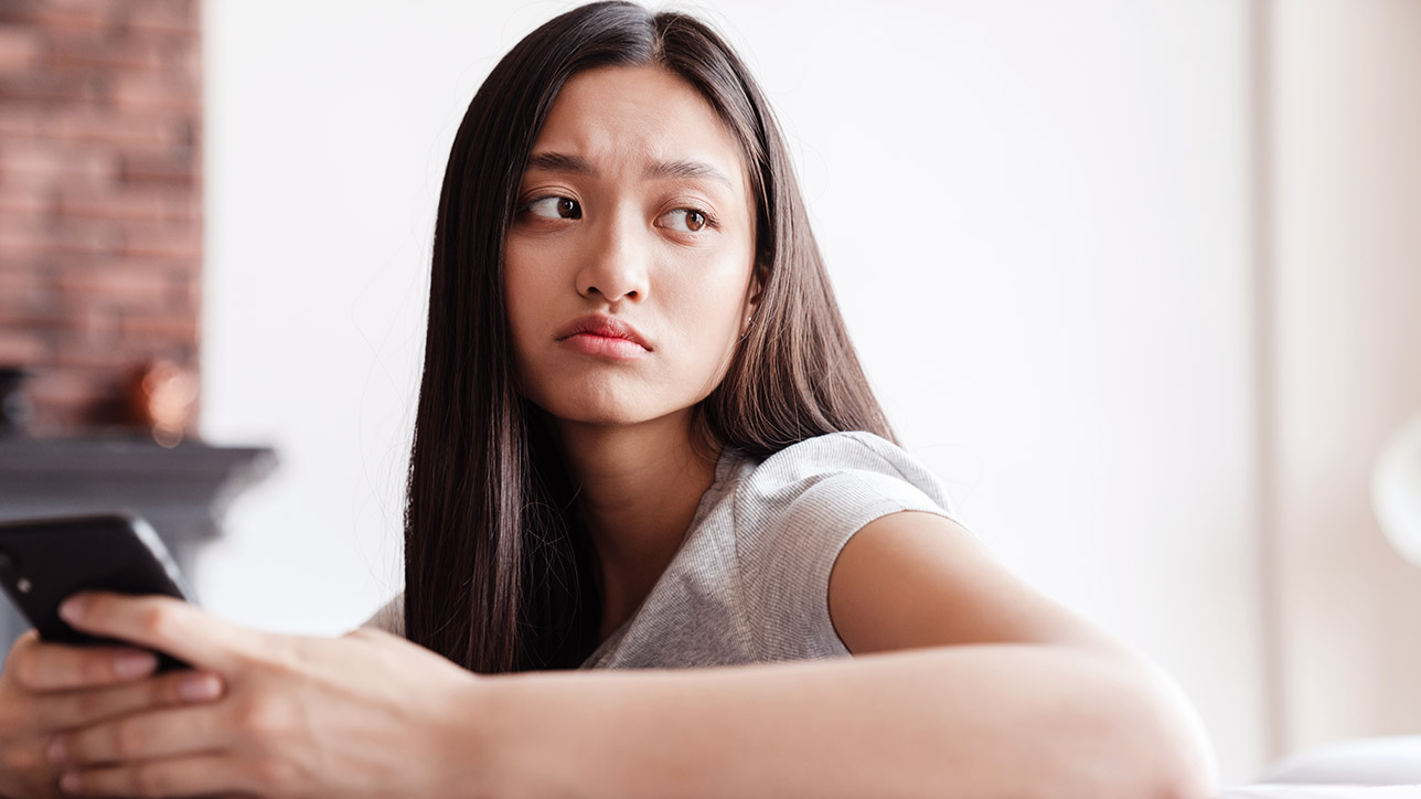 Image of an unhappy asian woman using a mobile phone while sitting on couch